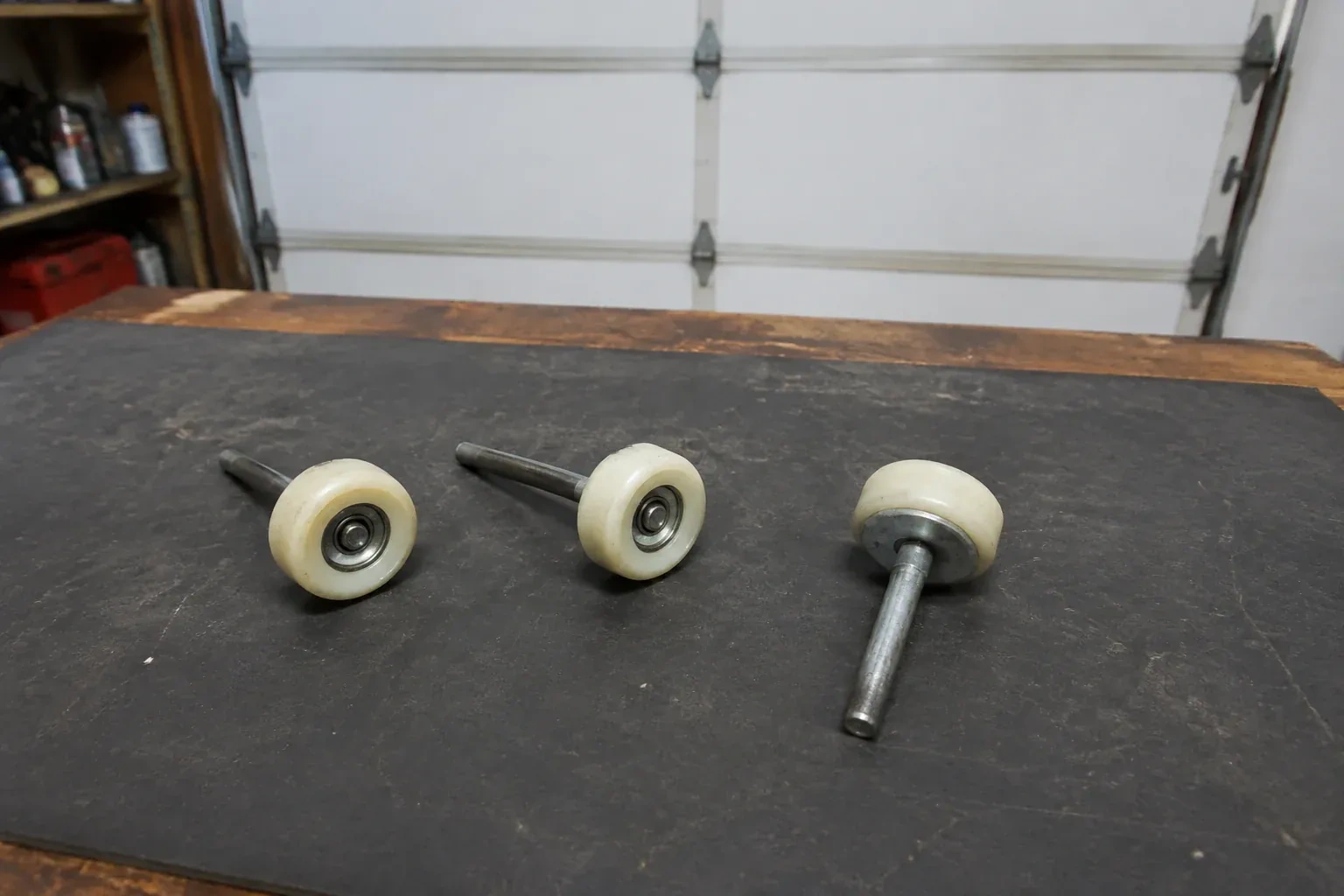 Three detached nylon-wheel garage door rollers with steel stems on a work mat. — Garage Door Roller Replacement, New Tecumseth